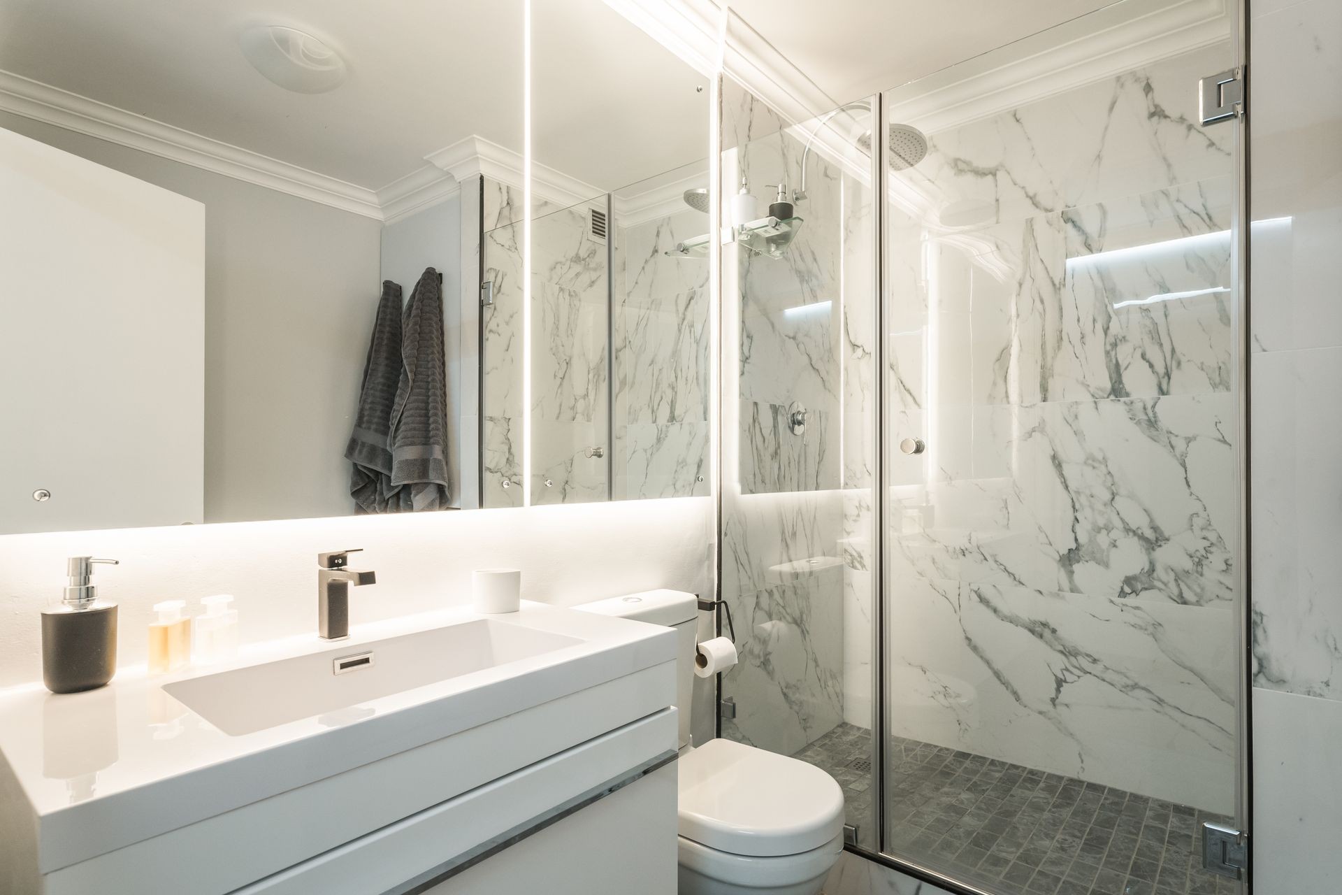 Modern, contemporary and brand new bathroom with marble walls and floor, white walls, cupboard and toilet, big back lit mirror